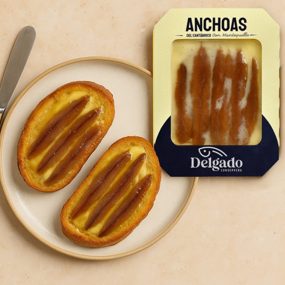 Two pieces of bread with anchovy spread on a plate next to a Delgado packaging.