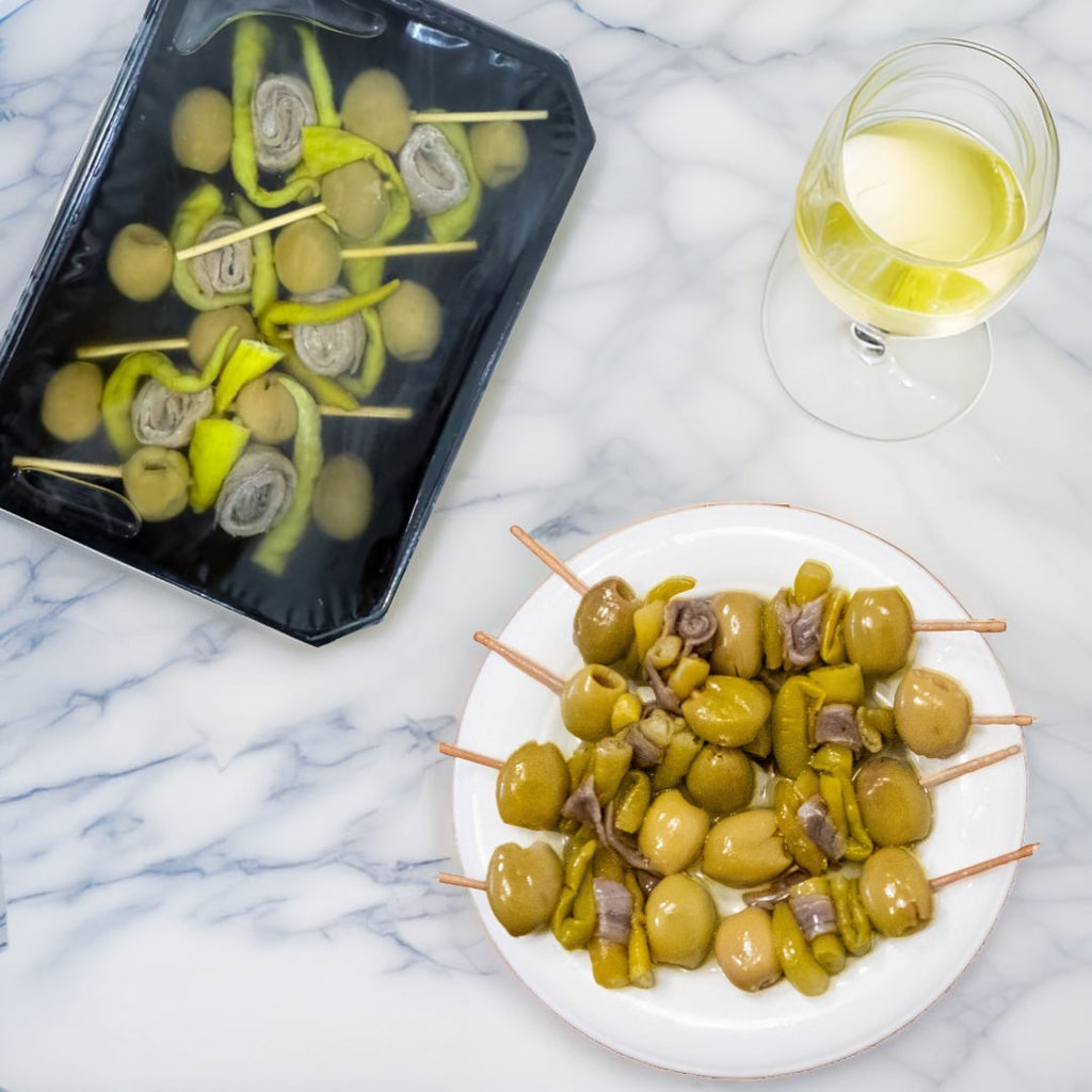 A plate of Cantabrian Anchovy Skewers Gildas with Manzanilla olives and guindilla peppers, served alongside a glass of white wine.