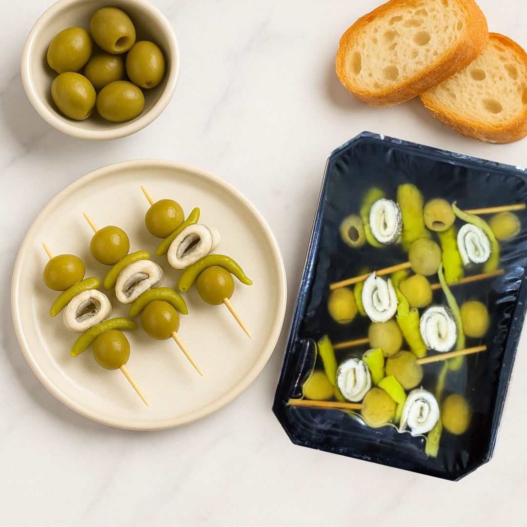 Boquerones skewers on a plate with a bowl of olives and slices of bread on a white surface.