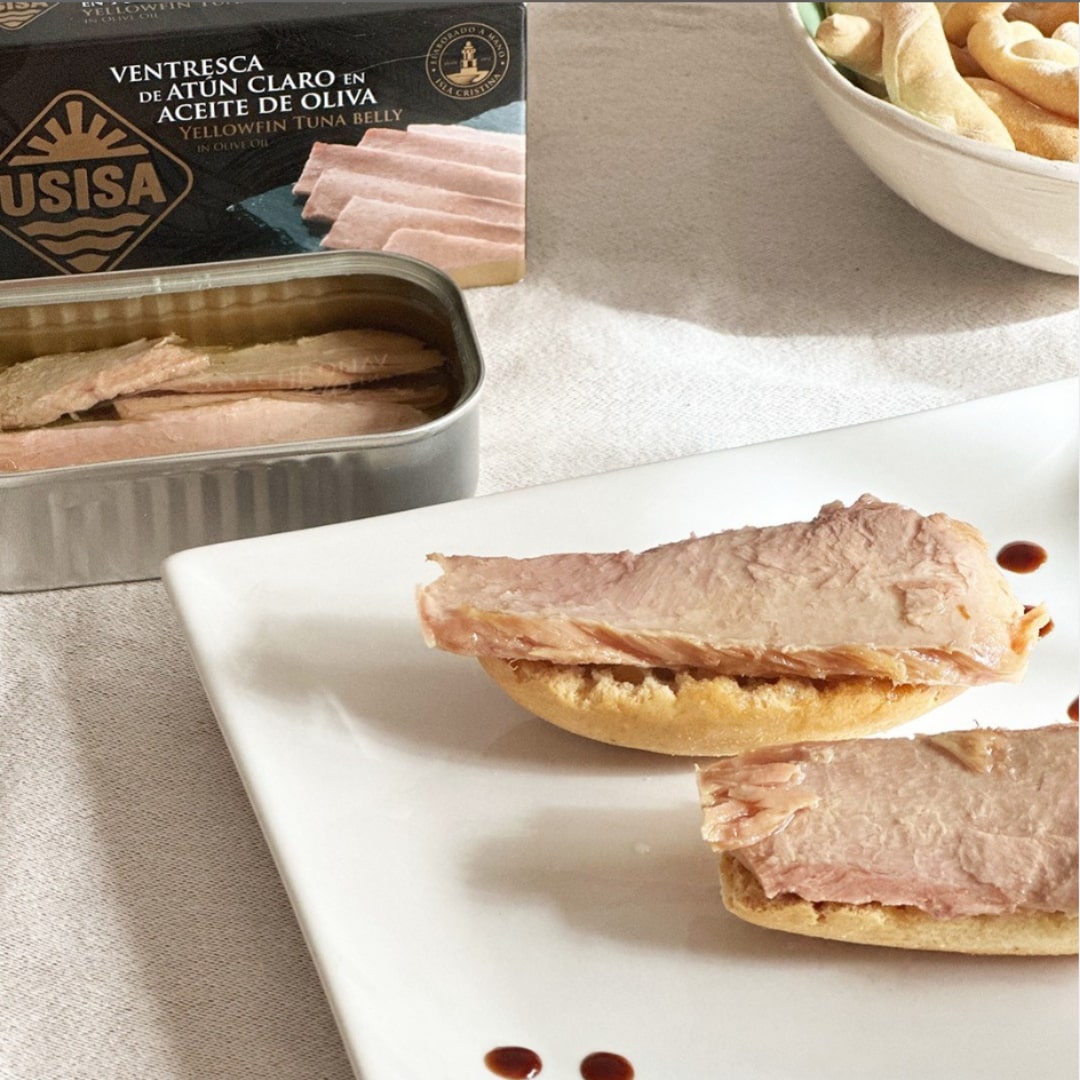 Canned tuna belly in olive oil by USISA, with slices of tuna on a toasted bread, and a bowl of olive oil in the background.