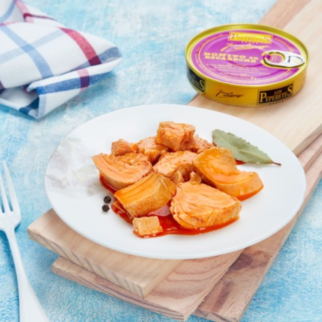 A plate of pickled white tuna, also known as Bonito Tuna in Escabeche, served on a white plate with a can of the product visible in the background. The tuna appears to be in a red pickled sauce, and there is a leaf of parsley garnish on the side.